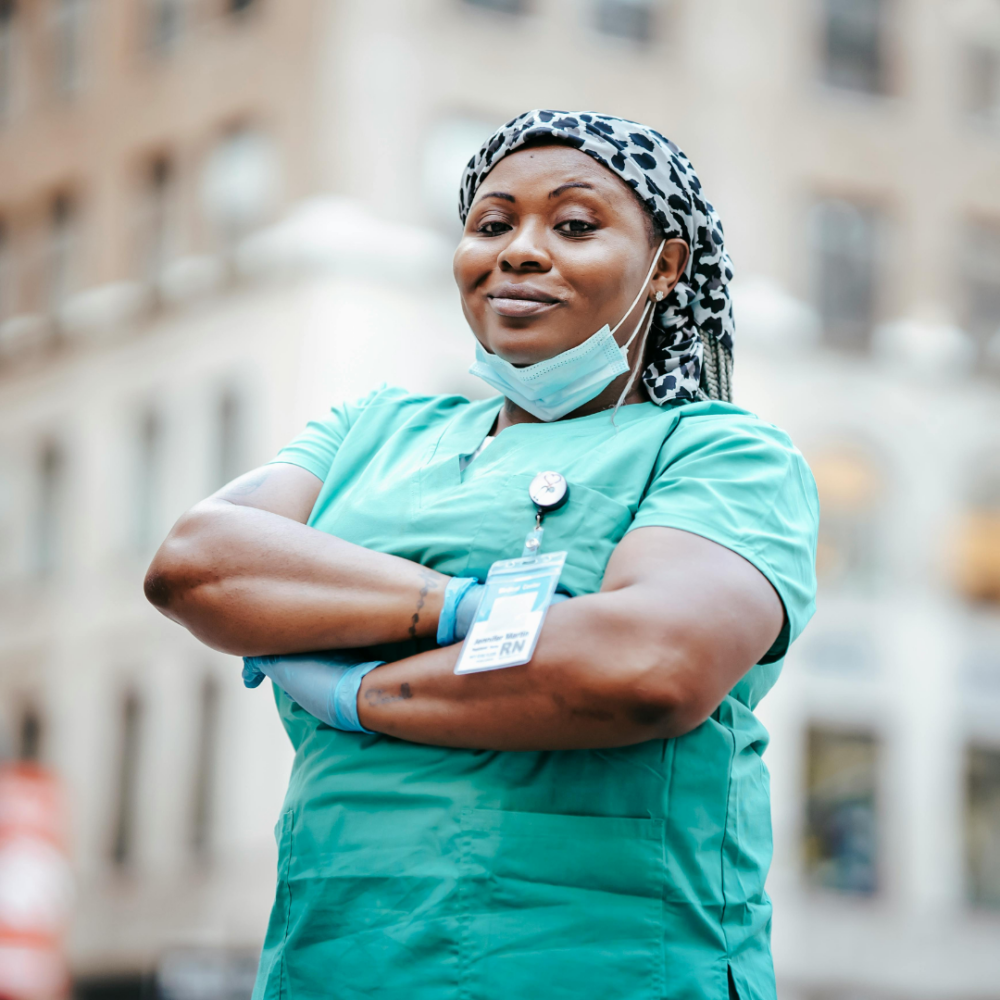 A nurse in green scrubs stands with arms crossed, wearing a patterned headscarf and a name badge, outside on an urban street. She has a face mask hanging loosely around her neck.